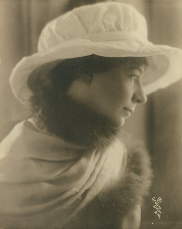 A sepia photograph of a woman wearing a large white hat and a jacket with a fur collar.