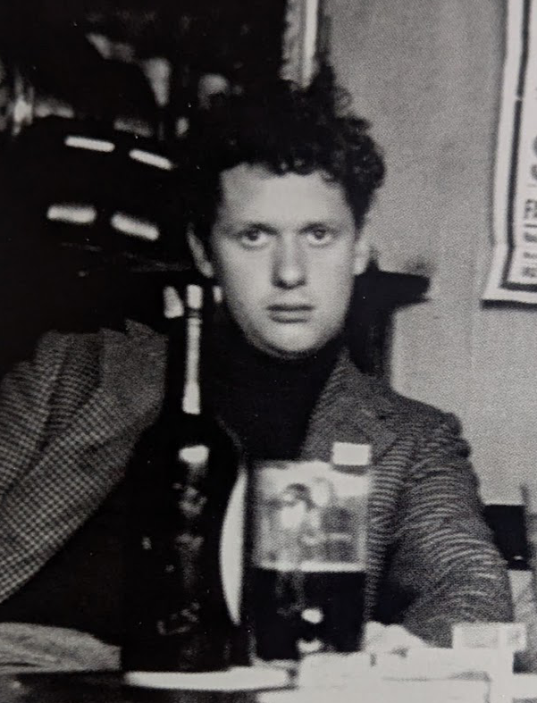 Black and white photograph of a young man sitting by a table. He has dark curly hair and wears a dark turtleneck shirt and a checkered jacket. 